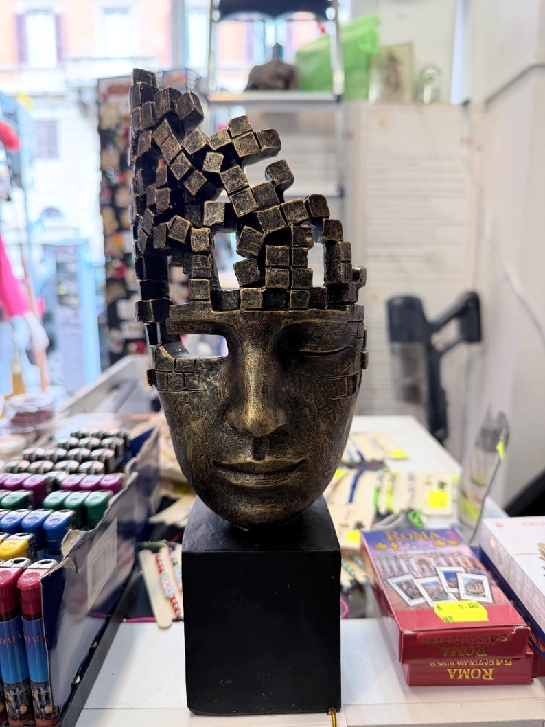 May include: A bronze-colored sculpture of a face with closed eyes and a fragmented top. The sculpture sits on a black rectangular base. The face has a serene expression, and the top of the head is made of small, square blocks.