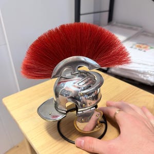 K&ouml;nnte beinhalten: Ein Miniaturhelm im r&ouml;mischen Stil aus poliertem, silberfarbenem Metall mit einem markanten, f&auml;cherf&ouml;rmigen Kamm aus leuchtend roten Borsten. Der Helm verf&uuml;gt &uuml;ber einen klappbaren Wangenschutz mit goldfarbenem Rand auf einem schwarzen St&auml;nder.