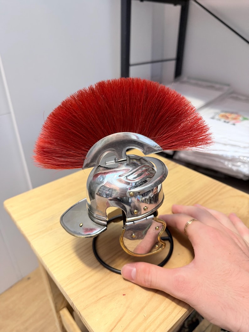 K&ouml;nnte beinhalten: Ein Miniaturhelm im r&ouml;mischen Stil aus poliertem, silberfarbenem Metall mit einem markanten, f&auml;cherf&ouml;rmigen Kamm aus leuchtend roten Borsten. Der Helm verf&uuml;gt &uuml;ber einen klappbaren Wangenschutz mit goldfarbenem Rand auf einem schwarzen St&auml;nder.