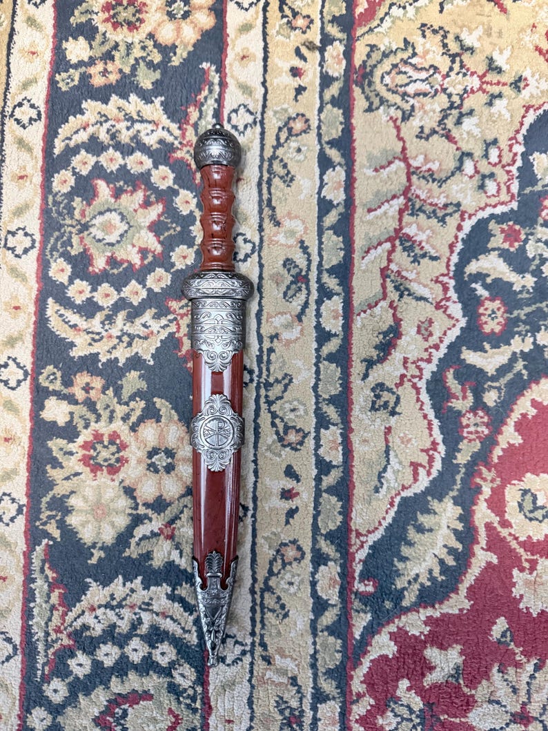 Pu&ograve; includere: Un pugnale racchiuso in un fodero di legno marrone con decorazioni in metallo color argento. L'impugnatura &egrave; in legno marrone con un pomolo arrotondato color argento. Il fodero presenta intricate incisioni metalliche e un emblema centrale.