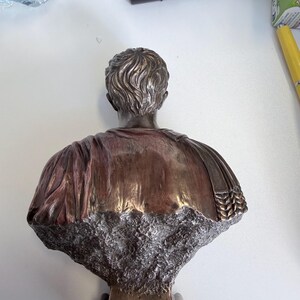 Peut inclure: Un buste de couleur bronze repr&eacute;sentant une personne vue de dos, avec des cheveux sculpt&eacute;s d&eacute;taill&eacute;s et des v&ecirc;tements drap&eacute;s. La partie inf&eacute;rieure de la sculpture pr&eacute;sente une finition rugueuse, textur&eacute;e et pierreuse qui contraste avec la surface m&eacute;tallique lisse.