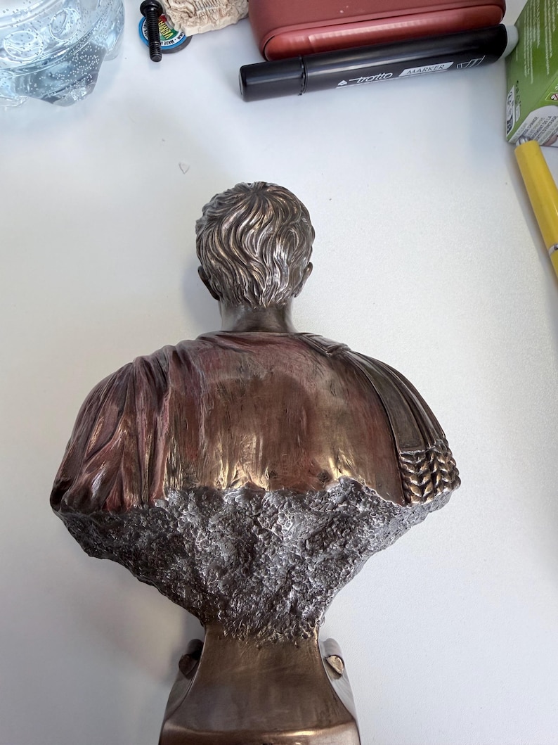 Peut inclure: Un buste de couleur bronze repr&eacute;sentant une personne vue de dos, avec des cheveux sculpt&eacute;s d&eacute;taill&eacute;s et des v&ecirc;tements drap&eacute;s. La partie inf&eacute;rieure de la sculpture pr&eacute;sente une finition rugueuse, textur&eacute;e et pierreuse qui contraste avec la surface m&eacute;tallique lisse.