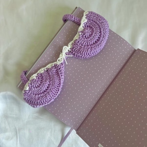 May include: A handmade, crochet bookmark in the shape of a bra, in a soft lavender color with white trim. The bookmark is resting on a lavender notebook with white polka dots. A white ribbon is tied between the bra cups.