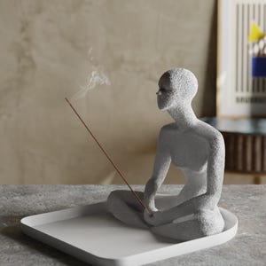 May include: A textured light grey sculpture of a seated figure in a meditative pose, positioned on a smooth white square tray. A thin brown incense stick is held by the figure, with a small wisp of white smoke rising from the lit tip.