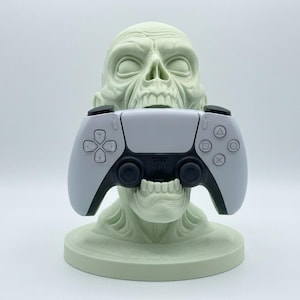 Könnte beinhalten: Ein hellgrüner, schädelartiger Gaming-Controller-Ständer, der einen weiß-schwarzen PlayStation 5-Controller hält. Der Ständer hat einen runden Sockel und detaillierte Gesichtszüge. Ein einzigartiges Accessoire für Gamer.