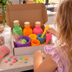 May include: A set of colorful silicone potion-making toys in a cardboard box, including three bottles with wooden stoppers, a purple cauldron, small cups, a yellow stirring spoon, a star-shaped wand, and small flower-shaped pieces in various colors.
