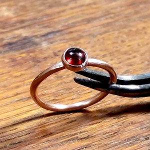 May include: A rose gold ring with a round, deep red gemstone. The ring is held by a pair of black tweezers against a wooden surface. The gemstone is approximately 0.25 inches in diameter.