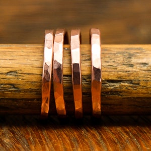 May include: Four hammered copper rings stacked on a wooden surface. The rings are a warm, reddish-brown color and have a textured, hammered finish.