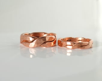 May include: Two copper braided rings, one larger than the other, with a hammered finish. The rings are on a white surface.