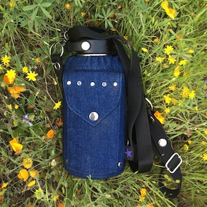 Pode incluir: Um porta-garrafa de ganga azul com rebites prateados e uma alça preta. A garrafa tem uma tampa preta e é rodeada por um campo de flores silvestres amarelas e roxas. O suporte tem uma aba com fecho de botão prateado.