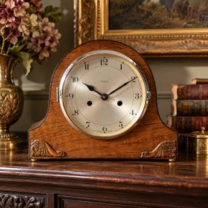 May include: An arched wooden mantel clock with a silver-toned circular face, black Arabic numerals, and black hands. The clock features the brand name Enfield printed on the dial, two winding holes, and decorative carved wooden corner accents.