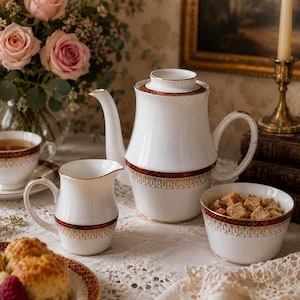 Könnte beinhalten: Ein weißes Porzellan-Teeservice mit Goldrand und rotem Band. Das Set beinhaltet eine Teekanne, einen Sahnekännchen, eine Zuckerdose und eine Teetasse. Ein Strauß rosa Rosen und Scones mit Himbeeren sind ebenfalls auf dem Tisch.