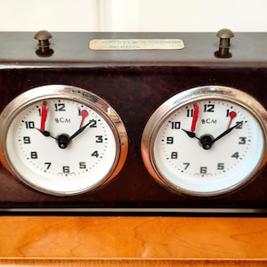 Puede incluir: Un reloj de ajedrez rectangular de color marrón jaspeado con dos esferas circulares blancas, números negros y la marca BCM. Cada esfera tiene manecillas negras para horas y minutos, y un segundero rojo. Dos botones de color latón en la parte superior.