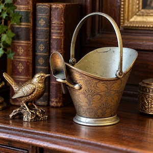 May include: A brass coal scuttle with a swing handle and a small attached shovel, featuring an etched floral pattern on the exterior. Beside it sits a small, detailed brass bird figurine perched on a textured base, both with a metallic finish.