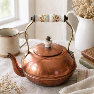Könnte beinhalten: Ein Kupferkessel mit rundem Körper und gebogenem Ausguss. Der Griff ist aus Messing und hat einen weißen Keramikgriff, der mit orangefarbenen und gelben Blumendesigns verziert ist. Ein passender Keramikknopf sitzt auf dem Deckel.
