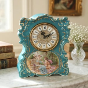 May include: A teal ceramic mantel clock with gold-painted scrollwork edges. The clock face features black Roman numerals and ornate hands. Below the clock face is an oval-shaped painted scene depicting a person and a cherub in a garden setting.