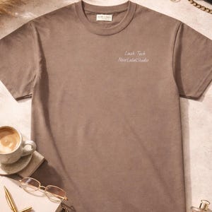 May include: A brown crew neck t-shirt with the text "Lash Tech NoirLabelStudio" in white. The shirt is displayed with accessories including a cup of coffee, glasses, and a perfume bottle.