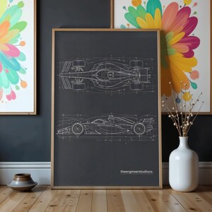 May include: A framed poster featuring a dark gray background with white technical line drawings of a Formula 1 race car in both top and side profiles. The text theengineerstudioco is printed in white at the bottom right of the poster.