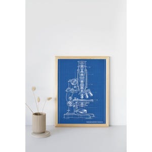 May include: A framed blueprint art print featuring a white line drawing of a microscope on a blue grid background. The print includes the text THEENGINEERSTUDIOCO at the bottom right corner. The frame is made of light-colored wood.