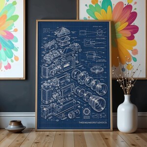 Könnte beinhalten: Ein gerahmtes Blaupausen-Poster mit einer Explosionszeichnung einer Kamera, bestehend aus weißen technischen Linienzeichnungen auf dunkelblauem Grund. Der Druck zeigt detaillierte mechanische Bauteile, Linsenelemente und den Text THEENGINEERSTUDIOCO.