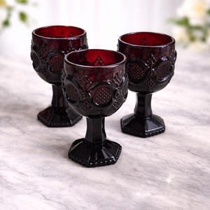 May include: Three ruby red wine glasses with ornate, embossed detailing. The glasses have a dark, hexagonal base and stem, and a round bowl. The glasses are arranged on a marble surface.