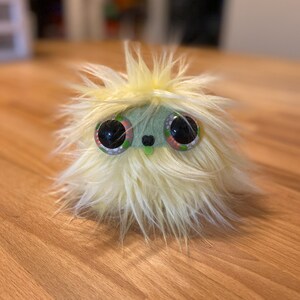 May include: A soft, fluffy toy creature with large eyes and a light green face. The creature has a yellow, fluffy exterior and a small, green nose. The eyes are black with green and red accents. The toy is on a wooden surface.
