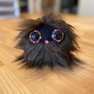 May include: A black furry toy with large pink and brown eyes and a pink tongue sticking out.