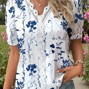 Peut inclure: Chemisier blanc à motif floral bleu. Le chemisier à col en V est orné d'une bordure en dentelle et de manches courtes bouffantes. Le motif floral présente des feuilles et des fleurs stylisées. Le chemisier est porté avec des colliers et des bracelets dorés.