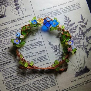 May include: A handcrafted floral crown featuring blue, yellow, and pink flowers, green leaves, and blue glass beads. The delicate wreath is set against a vintage book page background, creating a whimsical and artistic aesthetic.