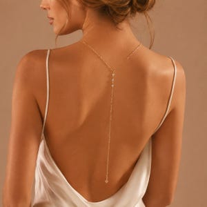 May include: A delicate gold-tone back necklace featuring a thin chain that drapes down the centre of the back. The necklace is adorned with small, clear, sparkling stones set at intervals along the upper portion of the chain, ending in a simple link.