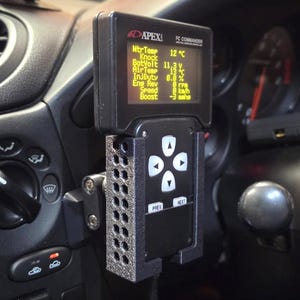 Può includere: Un APEXi FC Commander nero montato sul cruscotto di un'auto. Il display digitale mostra vari parametri del motore, come la temperatura dell'acqua e la pressione di sovralimentazione. L'unità ha pulsanti di controllo.