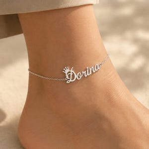 Peut inclure: Une chevillère argentée composée d'une chaîne fine et d'une plaque nominative personnalisée portant le nom Dorina en écriture cursive, avec une petite icône de couronne intégrée à la première lettre du nom, portée sur une cheville.