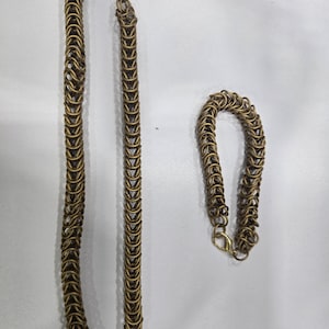 May include: A necklace and bracelet set made of interlocking bronze-colored metal rings. The necklace is long and hangs vertically, while the bracelet is a smaller, curved version. Both pieces have a clasp.