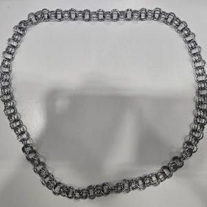 Puede incluir: Un collar plateado con un diseño de cota de malla. El collar está compuesto por eslabones circulares entrelazados, creando una apariencia flexible y texturizada. La cadena está colocada sobre una superficie blanca.
