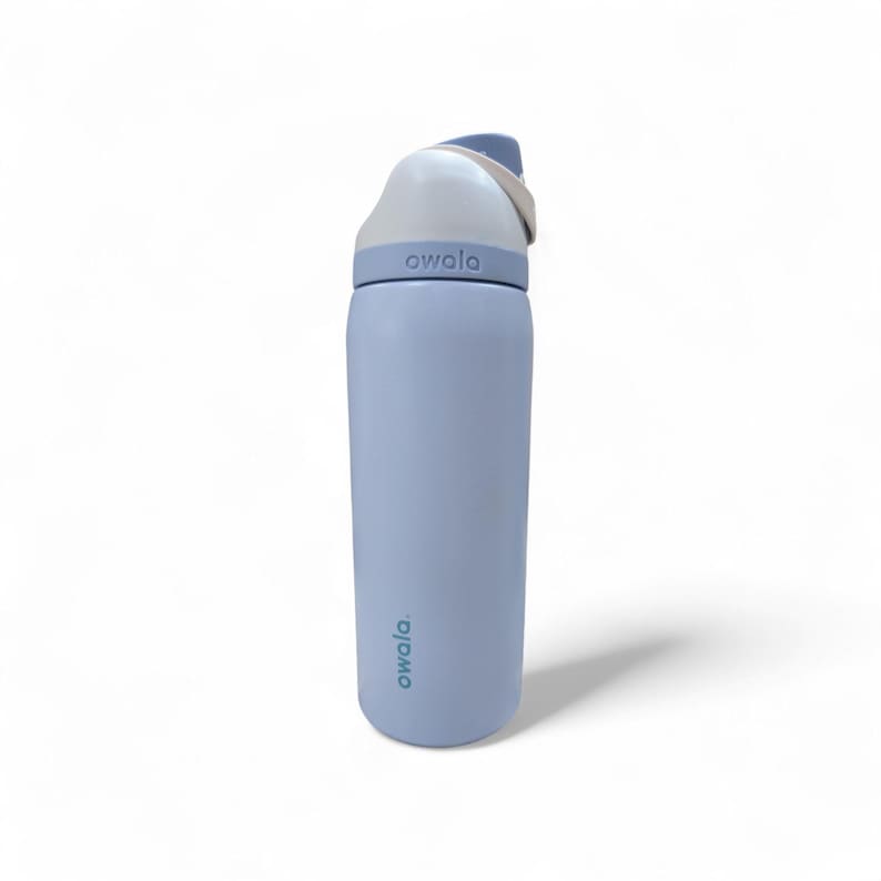 May include: A light blue Owala water bottle with a cylindrical body and a white lid featuring a carry loop. The brand name Owala is printed in dark text on the lower body and embossed on the lid collar. The bottle has a smooth matte finish.