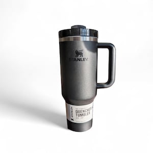 May include: A black Stanley Quencher tumbler with a handle and a black lid. The tumbler has the Stanley logo and the words "Quencher Tumbler" printed on a white label. The tumbler is designed to keep drinks cold all day.