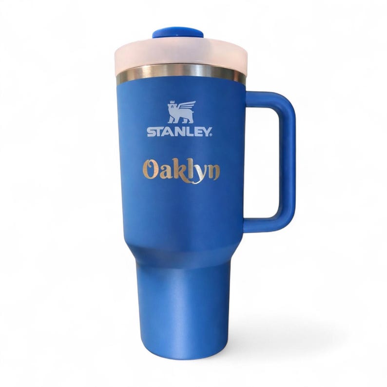 Może przedstawiać: Niebieski kubek podr&oacute;żny Stanley z uchwytem, białą obw&oacute;dką i niebieską pokrywką. Kubek ma złoty napis "Oaklyn" i logo Stanley. Kubek jest przeznaczony do utrzymywania ciepłych lub zimnych napoj&oacute;w.