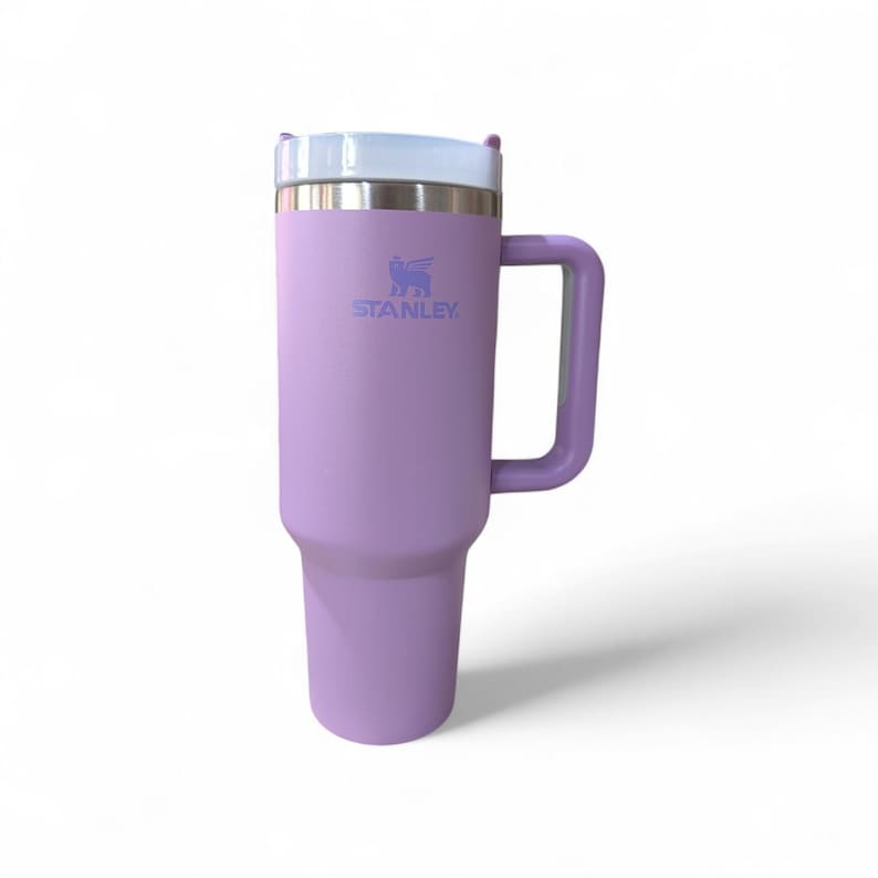 May include: A lavender Stanley tumbler with a handle. The tumbler has a white lid and a silver rim. The Stanley logo is printed in blue on the front. The tumbler is designed to keep drinks cold or hot for hours.