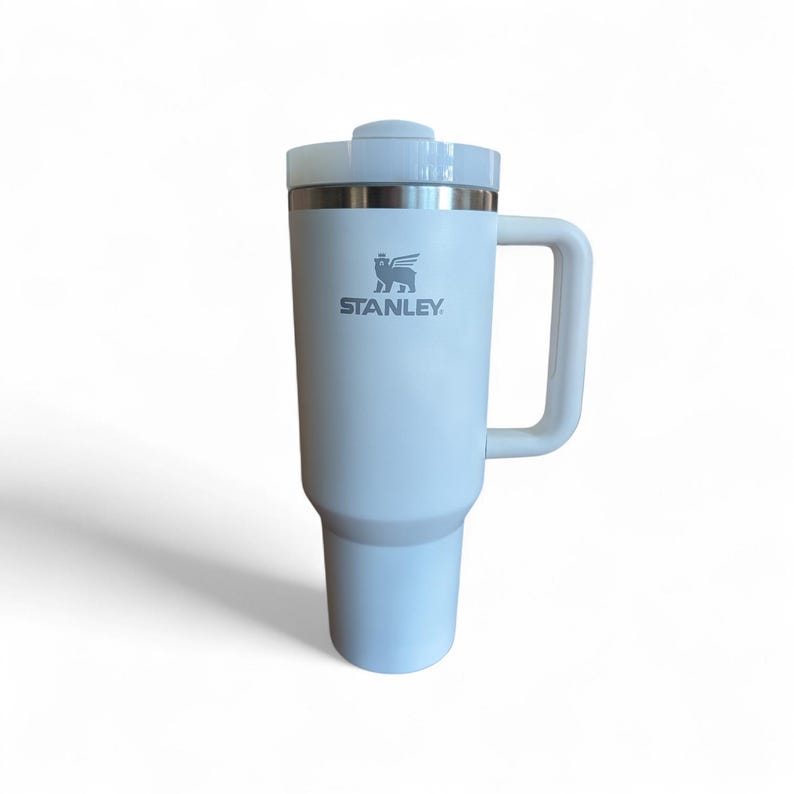 May include: A white Stanley travel mug with a handle and a lid. The mug has a cylindrical body with a slightly tapered base. The Stanley logo is printed in gray on the front. The mug is designed to keep beverages hot or cold.