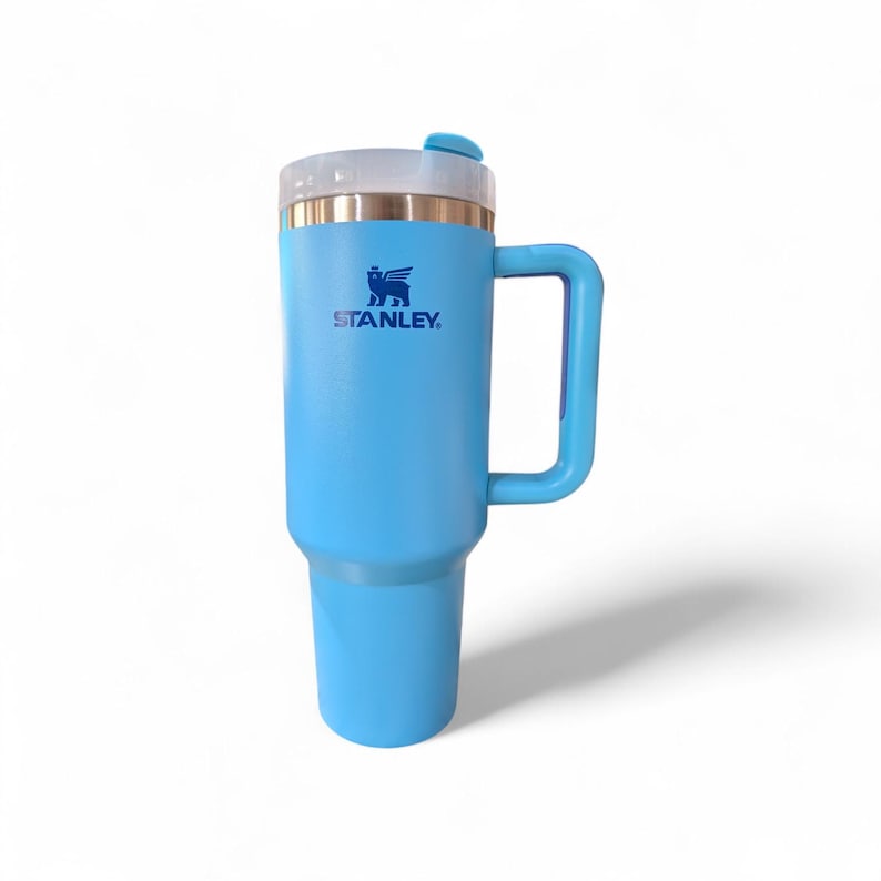 May include: A light blue Stanley travel mug with a handle. The mug has a clear lid and a gold-colored rim. The Stanley logo is printed in blue on the front of the mug.