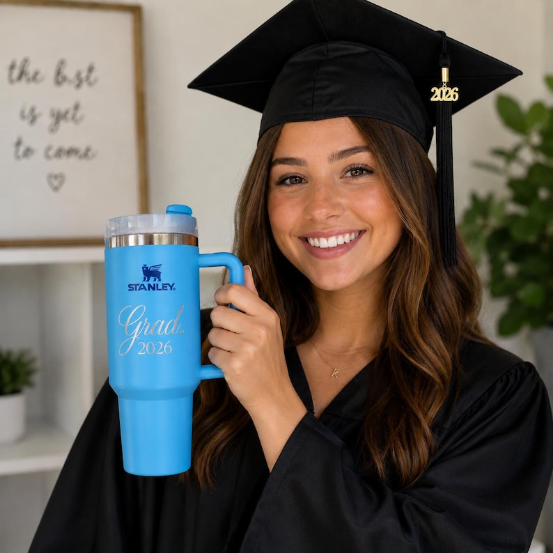 May include: A light blue Stanley tumbler with a handle, featuring the text "Grad 2026" in a script font. The tumbler has a silver lid and a black handle. The Stanley logo is visible. The tumbler is held by a person wearing a graduation cap.
