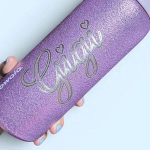 May include: A purple glittery Owala water bottle with a light purple cap. The bottle features the name Gigi engraved in a cursive script font with two small heart icons above the letters. The brand name Owala is printed on the side of the bottle.
