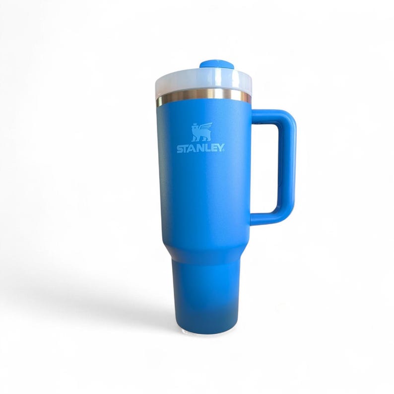May include: A blue Stanley travel mug with a handle and a white and gold lid. The Stanley logo is visible on the side. The mug is designed to keep beverages hot or cold. The mug is approximately 9 inches tall.