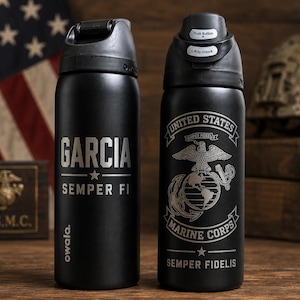May include: Two matte black Owala water bottles with black lids. The left bottle features the name GARCIA and SEMPER FI. The right bottle displays the United States Marine Corps emblem, the text UNITED STATES MARINE CORPS, and SEMPER FIDELIS.