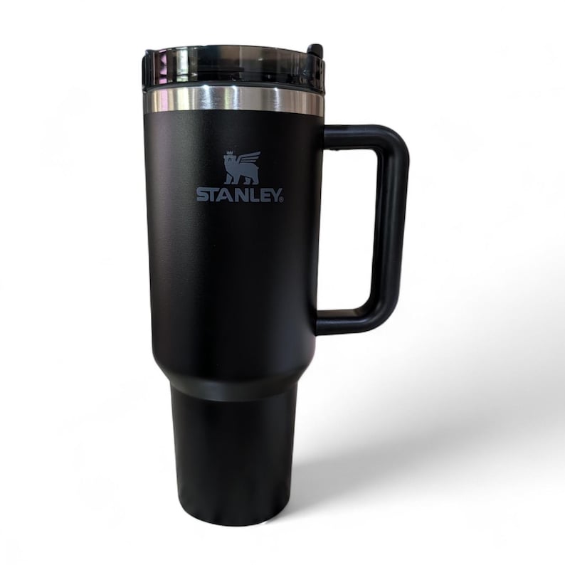 May include: Black Stanley travel mug with a handle and a clear lid. The Stanley logo is printed in white on the front of the mug. The mug is designed to keep beverages hot or cold for hours.