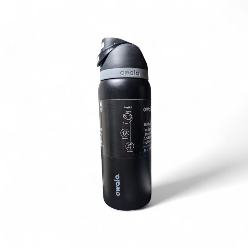 May include: A matte black 32 oz Owala water bottle with a gray lid and a black carry loop. The bottle features a black product label with white text and diagrams detailing the FreeSip spout, including the text Hi there, I'm leak-free, I'm insulated.