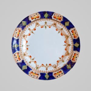 Op de afbeelding: Een ronde, witte keramische plaat met een gedetailleerd ontwerp. De rand heeft een diepblauwe rand met bloemen- en geometrische patronen in oranje, geel en groen. Het midden van de plaat is effen wit, wat contrasteert met de sierlijke rand.