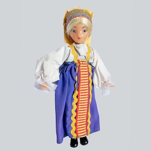 Könnte beinhalten: Eine Vintage-Puppe in traditioneller Kleidung. Die Puppe hat blondes Haar und blaue Augen und trägt eine weiße Bluse mit Spitzenmanschetten, einen blauen Rock und eine gelb-rote Zierblende. Der Kopf der Puppe ist mit einem gelben Schal und einem dekorativen Stirnband bedeckt.