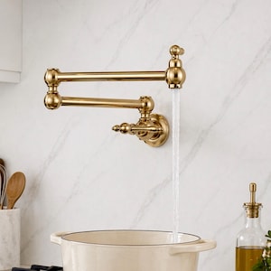 Puede incluir: Un grifo de llenado de ollas dorado, montado en la pared, con un brazo articulado. El agua fluye del grifo a una olla blanca. El grifo tiene un diseño clásico y está montado en una pared blanca con vetas de mármol.
