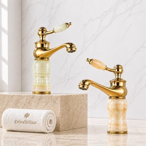 May include: Two bathroom taps with polished gold metal finishes and stone bases. One tap features a pale green stone base and handle, while the other has a beige marbled stone base and handle. A white towel with OrivelleHaus text is present.